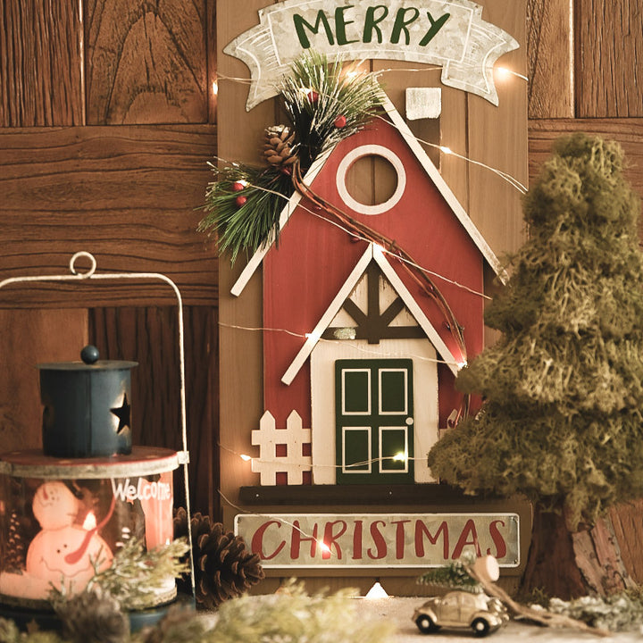 Decorative Christmas scene with a red barn-like structure, Christmas lights, and festive decorations on a wooden wall.