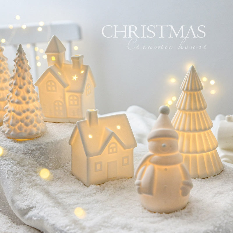 Ceramic Christmas decorations including houses, trees, and a snowman on a snowy surface with lights in the background.